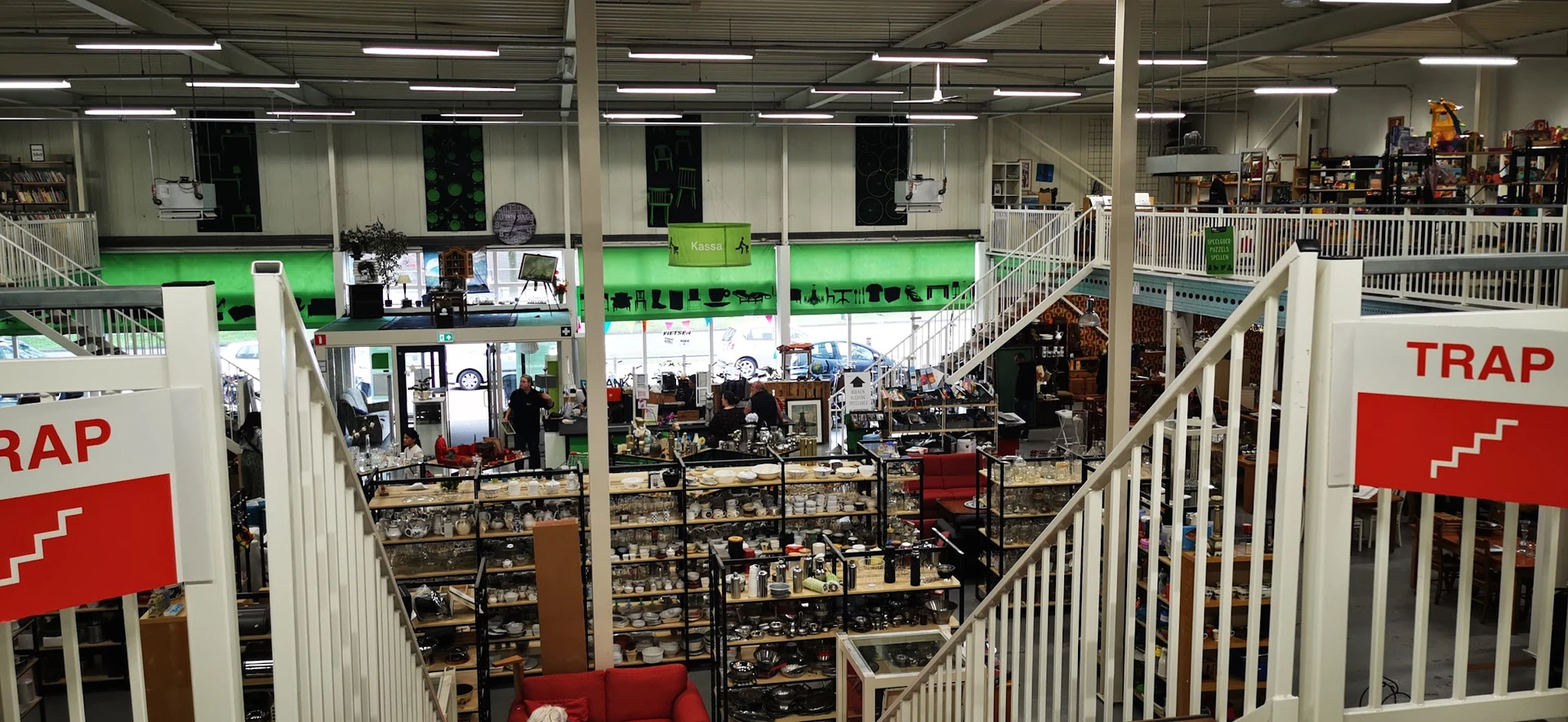 Overzicht van kringloopwinkel met meerdere verdiepingen, stellingen vol spullen, trappen en reling zichtbaar