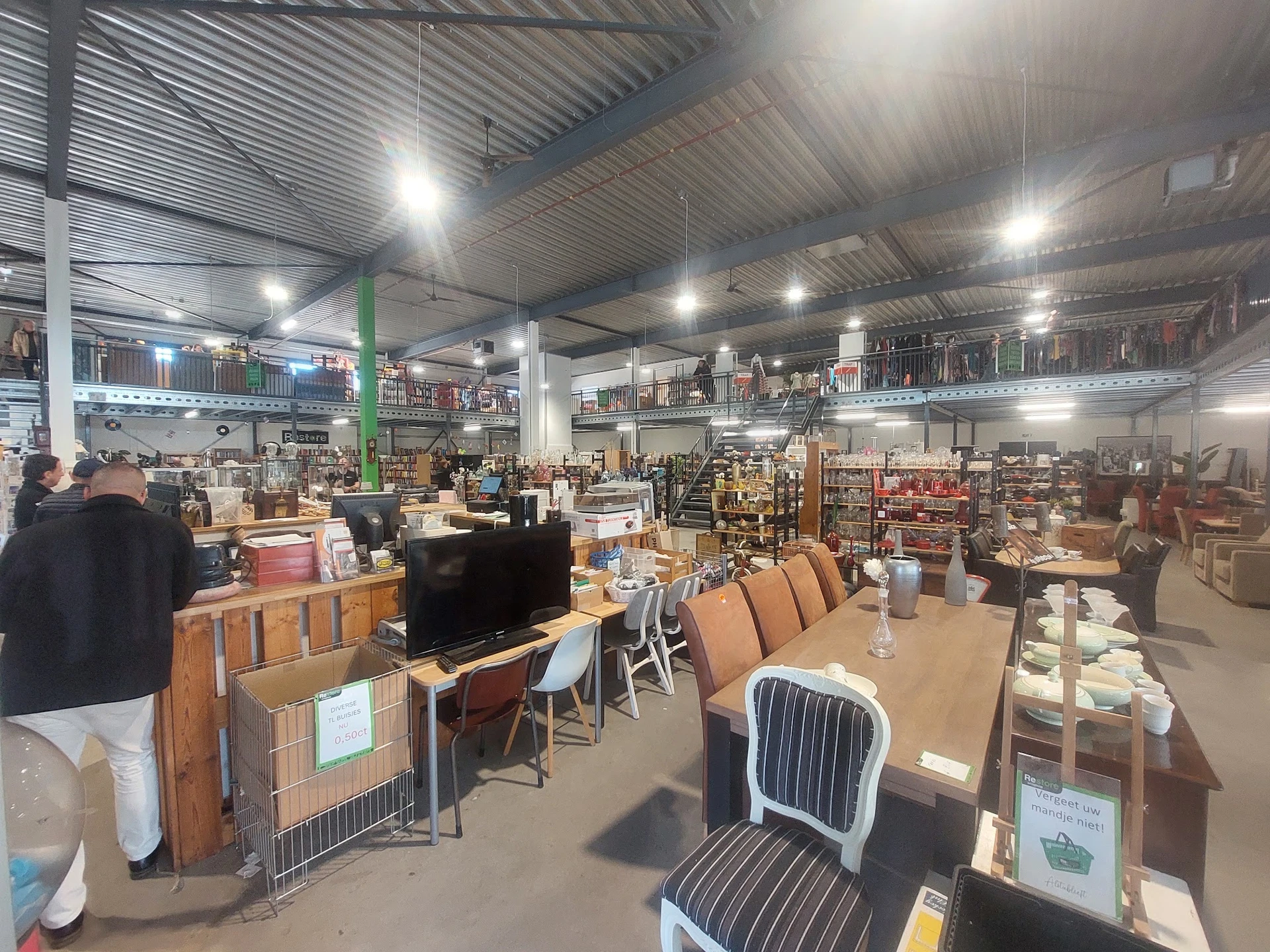 Overzicht van kringloopwinkel met meubels, stoelen, televisie en diverse spullen op de vloer en stellingen