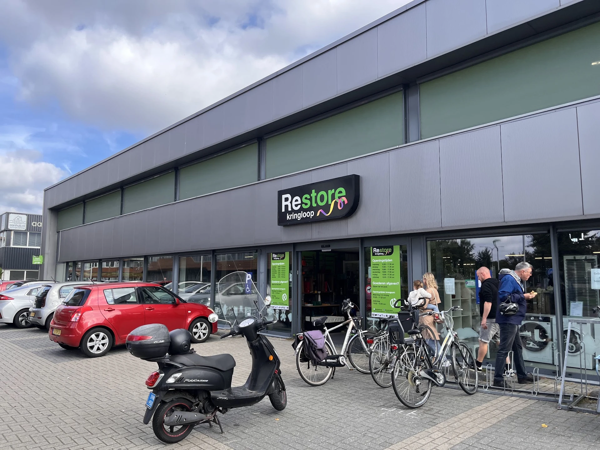 Gevel van Restore kringloopwinkel met logo, grote ramen, ingang, en parkeerplaats met auto's en fietsen ervoor