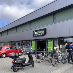 Gevel van Restore kringloopwinkel met logo, grote ramen, ingang, en parkeerplaats met auto's en fietsen ervoor
