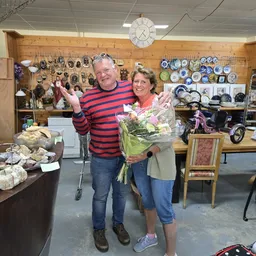 Man en vrouw poseren samen in een kringloopwinkel met bloemen, omgeven door servies en spullen