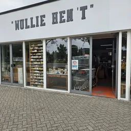 Gevel van kringloopwinkel met grote ramen, winkelnaam bovenaan, stellingen met spullen zichtbaar, ingang met oranje vloer