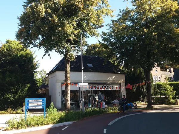 Witte kringloopwinkel aan de straatzijde met naam op gevel, grote ramen en ingang, omgeven door bomen