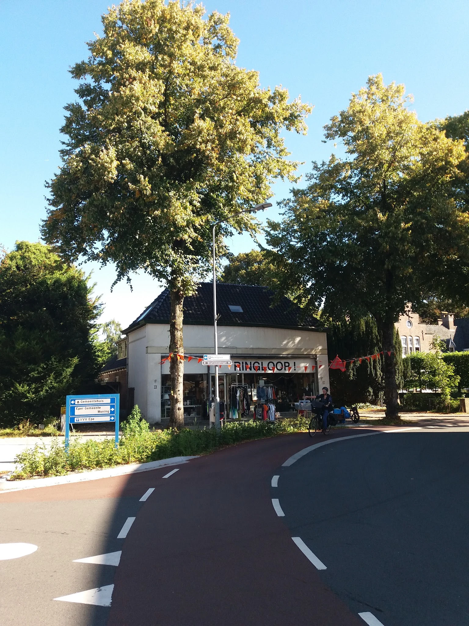 Witte kringloopwinkel aan de straatzijde met naam op gevel, grote ramen en ingang, omgeven door bomen