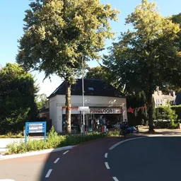 Witte kringloopwinkel aan de straatzijde met naam op gevel, grote ramen en ingang, omgeven door bomen