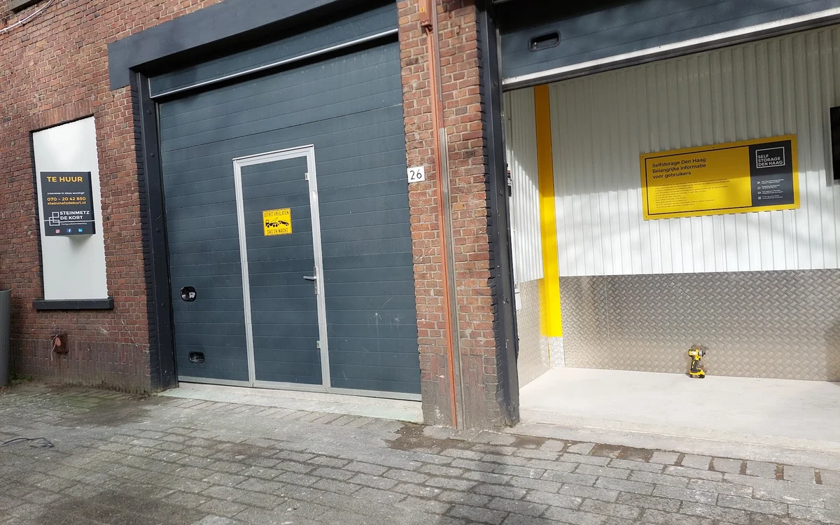 Grijze garagedeur en witte metalen deur met gele waarschuwingsstickers op bakstenen gevel