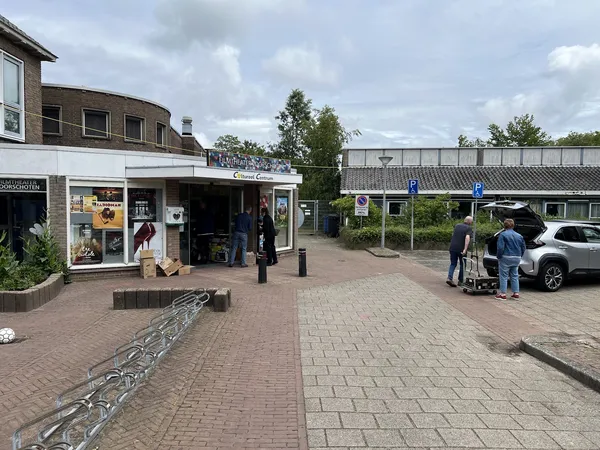 Voorgevel van kringloopwinkel met etalage, ingang en parkeerplaats met auto's en bezoekers