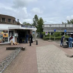 Voorgevel van kringloopwinkel met etalage, ingang en parkeerplaats met auto's en bezoekers