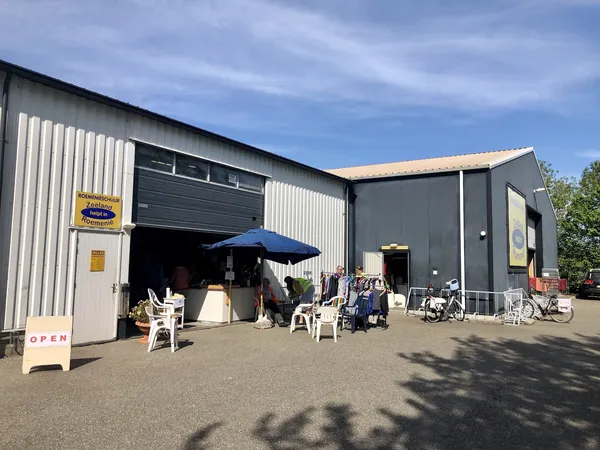 Exterieur van kringloopwinkel met grijze en witte metalen gevel, openstaande garagedeur, witte stoelen op terras onder blauwe