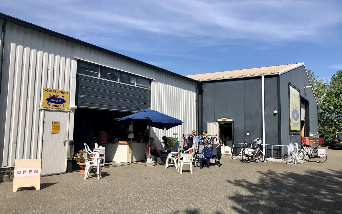Exterieur van kringloopwinkel met grijze en witte metalen gevel, openstaande garagedeur, witte stoelen op terras onder blauwe