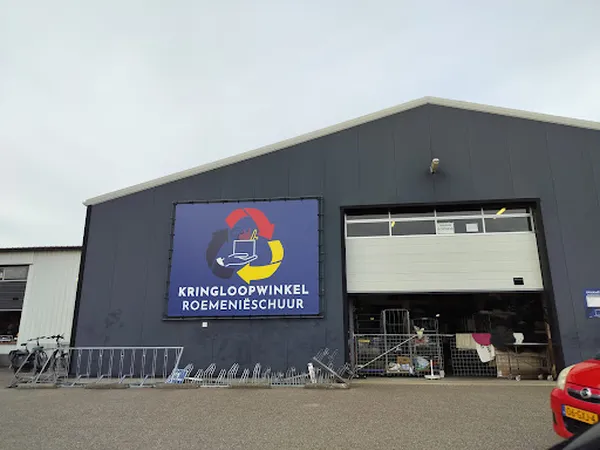 Grijze gevel van kringloopwinkel met blauw logo en grote garagedeur, winkeluit zichtbaar op parkeerterrein