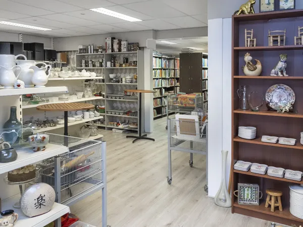 Overzicht van een kringloopwinkel met stellingen vol servies, boeken, decoratieve items en een donkerbruine vitrinekast met p