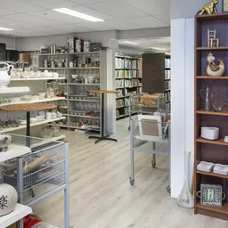 Overzicht van een kringloopwinkel met stellingen vol servies, boeken, decoratieve items en een donkerbruine vitrinekast met p