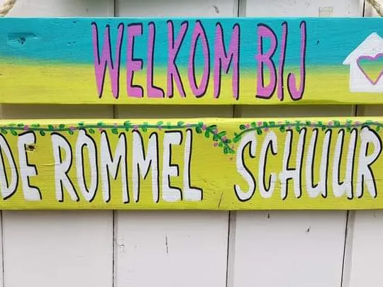 Welkomsbord met 'Welkom Bij' en 'De Rommel Schuur' in paars en geel op witte houten wand