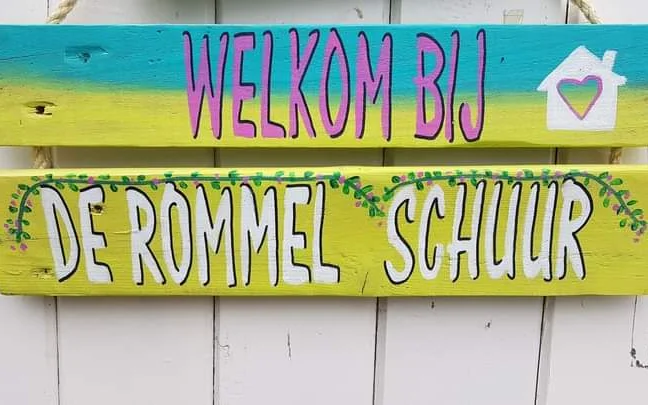 Welkom-uithangbord en winkelnaambord 'De Rommel Schuur' op witte houten wand