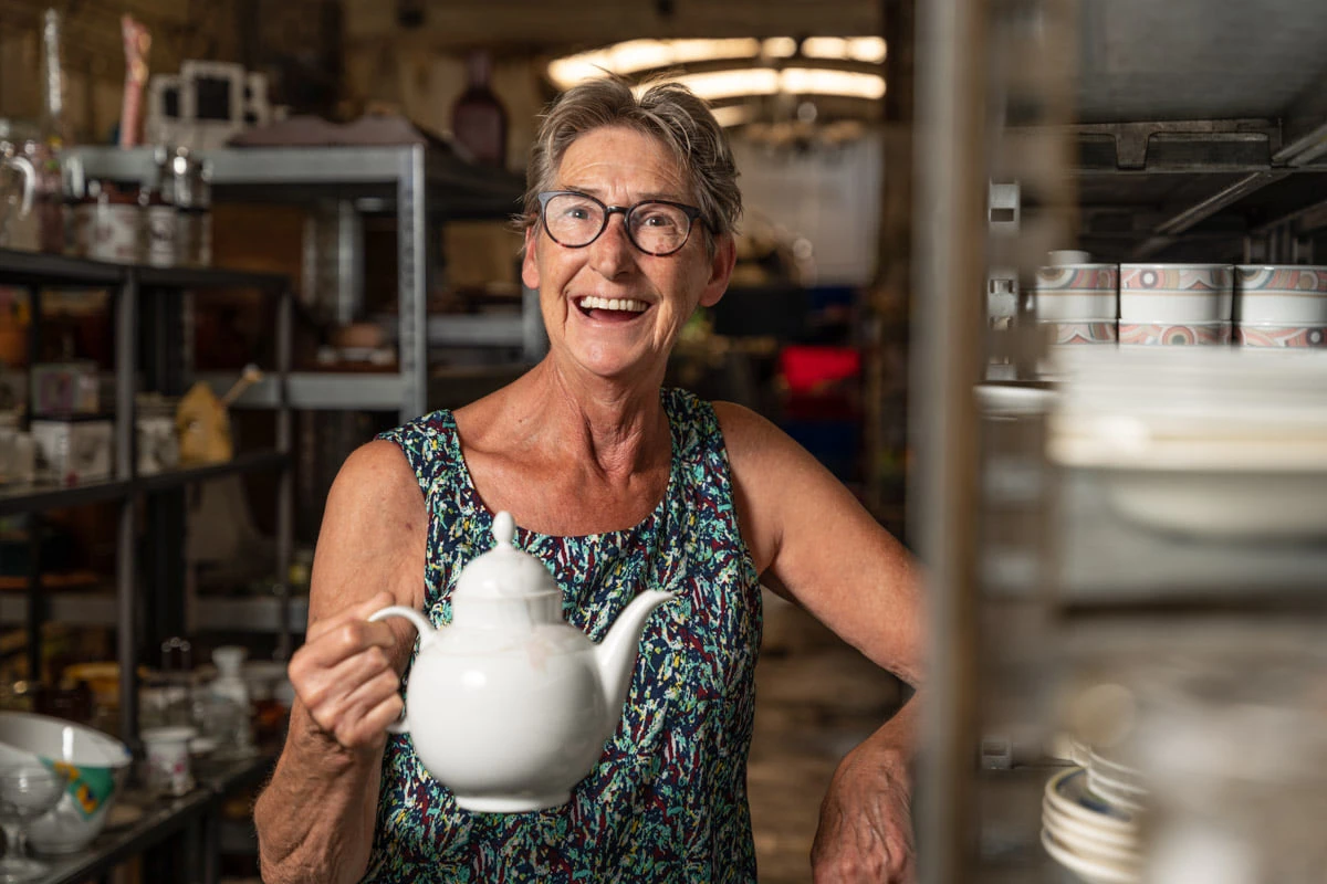 Vrouw met bril en gekleurde mouwloze top houdt witte theepot vast in kringloopwinkel