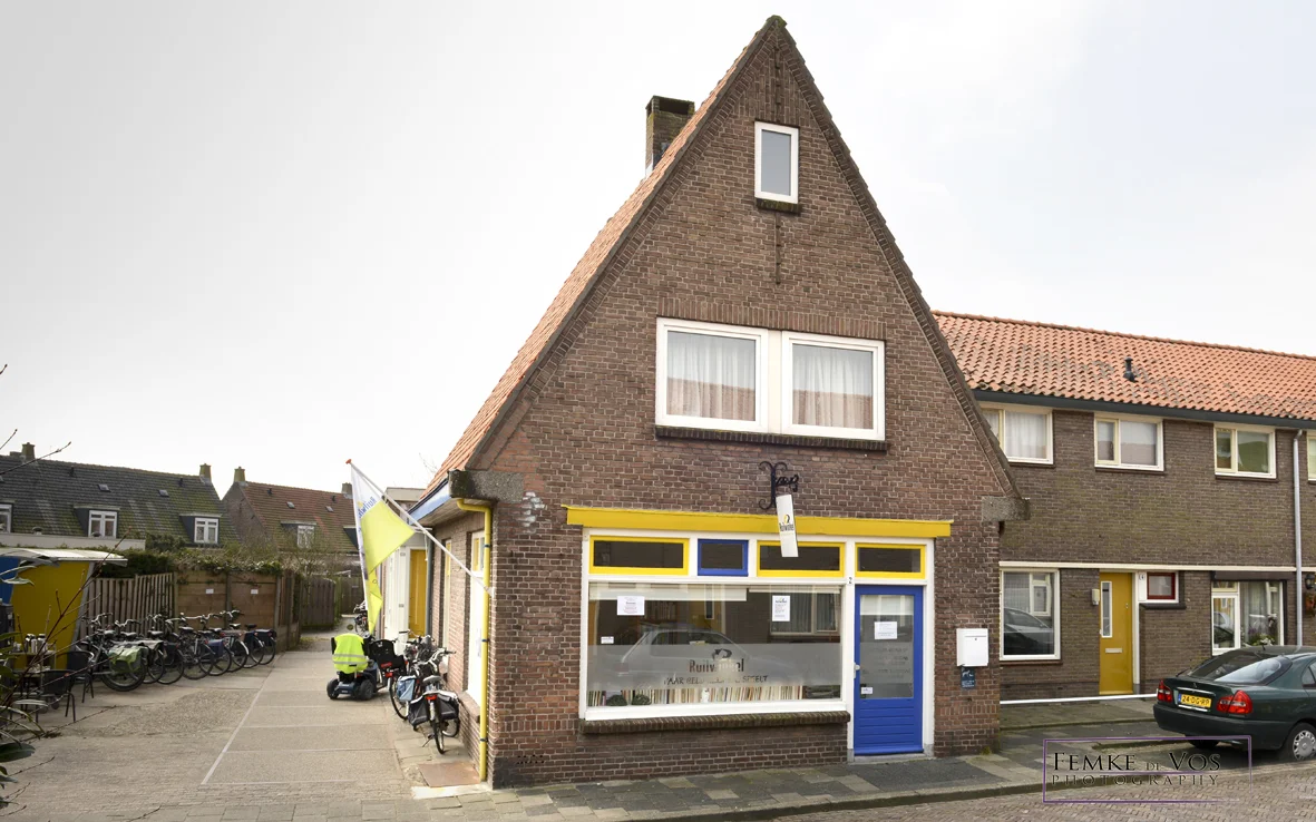 Bakstenen gevel van kringloopwinkel met gele en blauwe pui, etalageruit en ingangseur aan straat