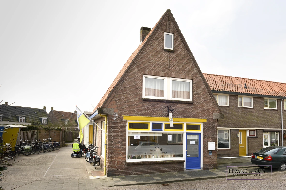 Bakstenen gevel van kringloopwinkel met gele en blauwe pui, etalageruit en ingangseur aan straat
