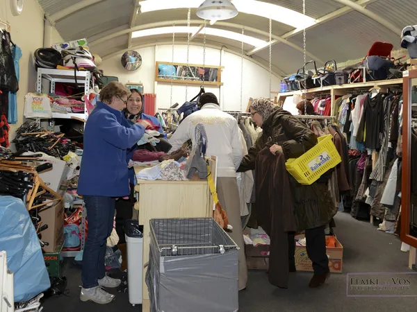 Klanten winkelen tussen rekken met kleding en dozen in een kringloopwinkel met daarbovenin zichtbare skylight.