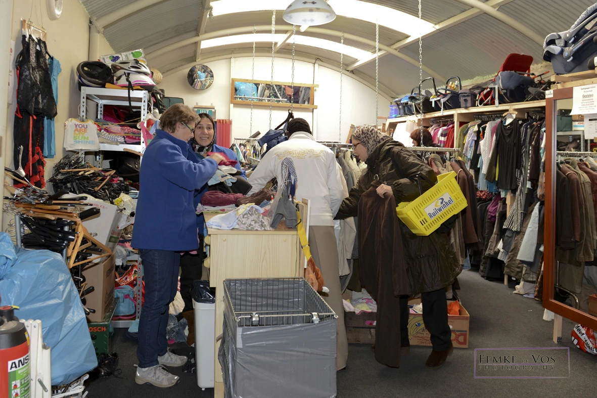 Klanten winkelen tussen rekken met kleding en dozen in een kringloopwinkel met daarbovenin zichtbare skylight.