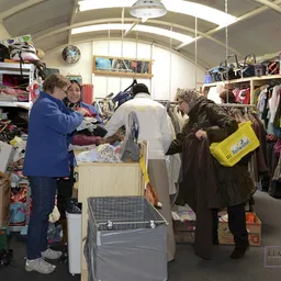 Klanten winkelen tussen rekken met kleding en dozen in een kringloopwinkel met daarbovenin zichtbare skylight.