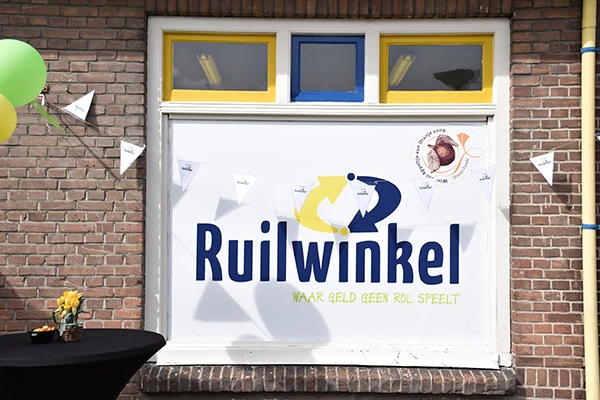 Gevel van kringloopwinkel Ruilwinkel met wit winkelfront en blauwe/gele logo, gele en blauwe ramen boven de deur