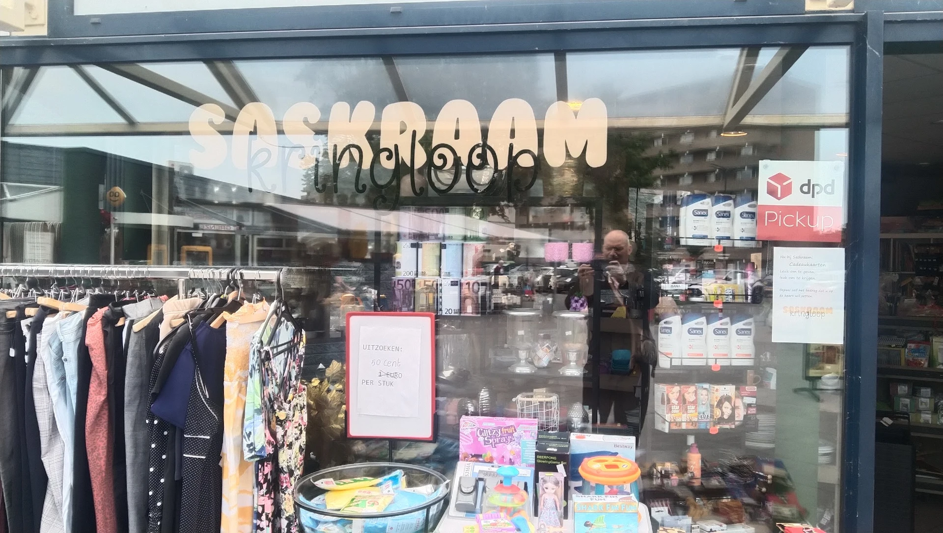 Winkelruit met naam 'saskraam' boven de deur, uitstalraam met kleding en diverse spullen zichtbaar