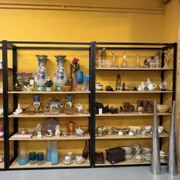 Twee zwarte stellingen vol met verzamelde voorwerpen zoals vazen, beeldjes, servies, flessen en decoratieve items tegen gele 