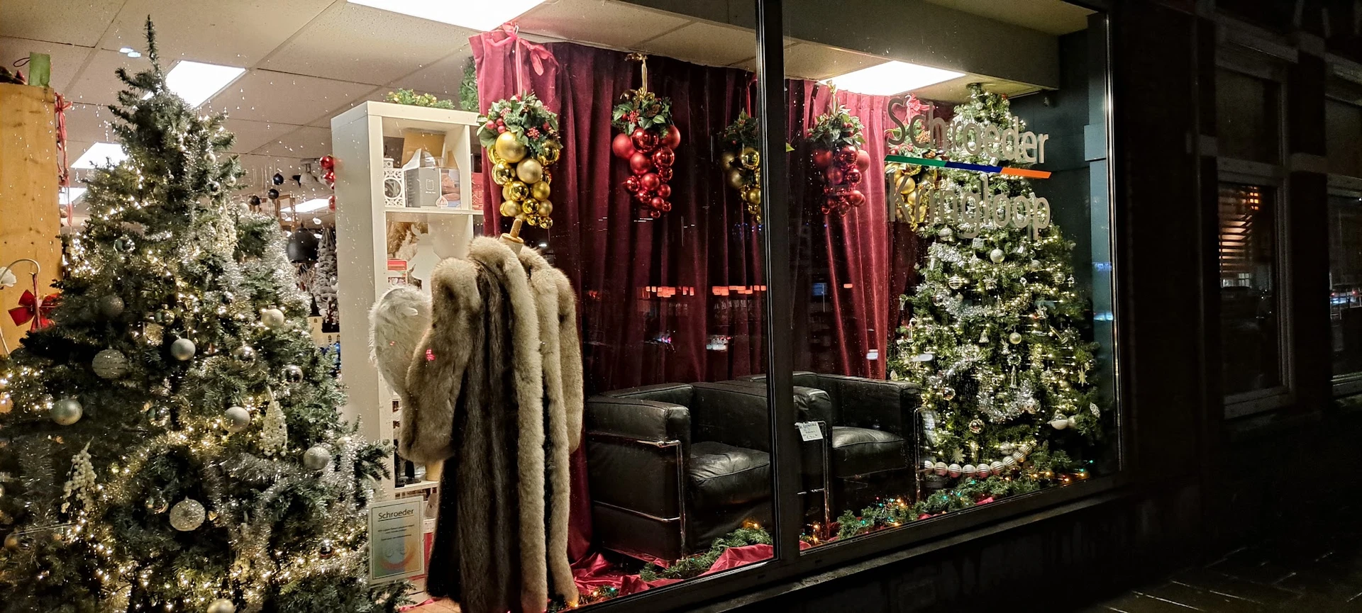 Etalage met kerstdecoratie: rode gordijnen, hangende bloemstukken, kerstbomen, zwarte fauteuils en een bontjas op etalagepop
