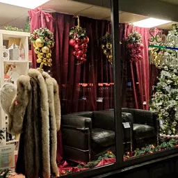 Etalage met kerstdecoratie: rode gordijnen, hangende bloemstukken, kerstbomen, zwarte fauteuils en een bontjas op etalagepop