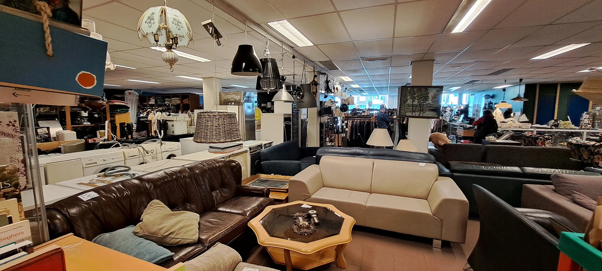 Winkelvloer met bruine leren bank, crèmekleurige zitbank en houten salontafel, achter winkelrekken met kleding en hangende ar