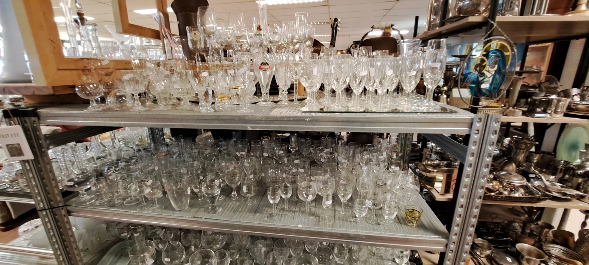 Glazen servies en drinkglazen uitgestald op metalen stellingen in een kringloopwinkel