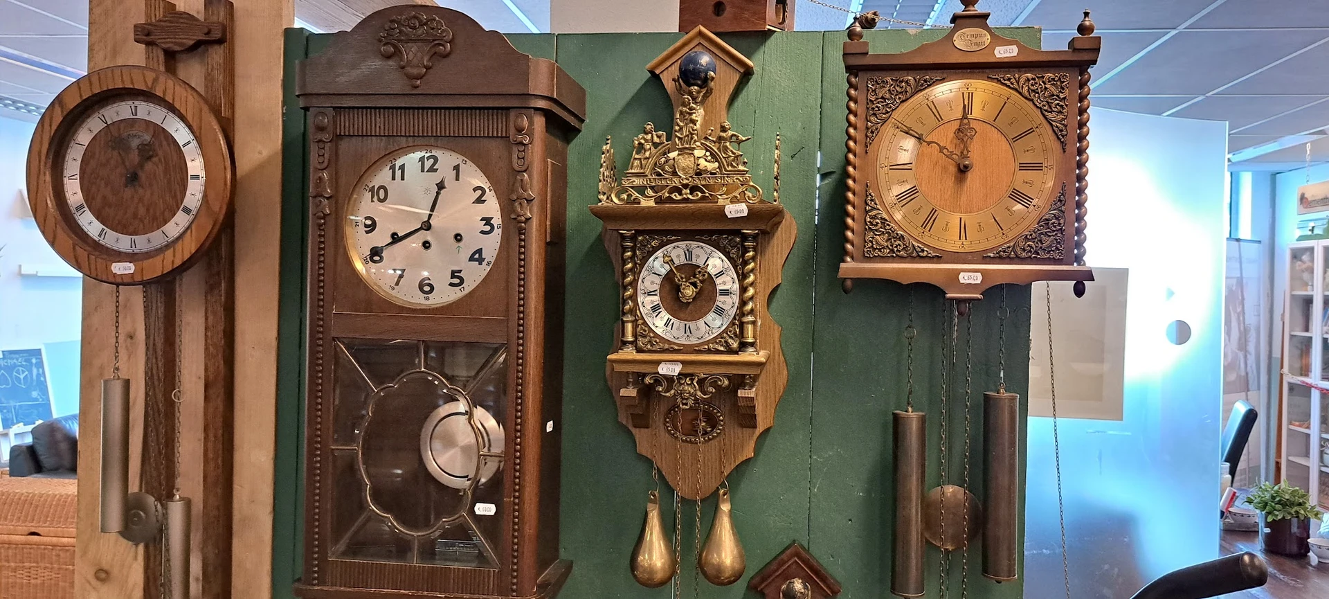 Drie antieke wandklokken met slingers uitgestald tegen groene wand in kringloopwinkel