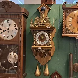 Drie antieke wandklokken met slingers uitgestald tegen groene wand in kringloopwinkel