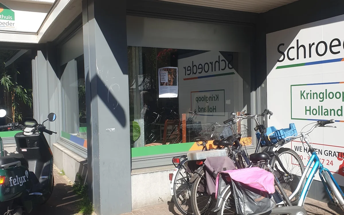 Voorkant van kringloopwinkel met vitrine, fietsen geparkeerd ervoor en winkelnaam gedeeltelijk zichtbaar