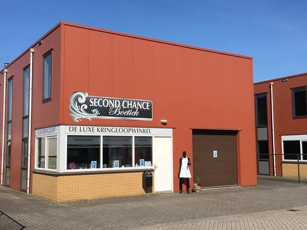 Rode bakstenen gevel van kringloopwinkel Second Chance Boetiek met witte winkelruit en bruine deur, persoon loopt langs
