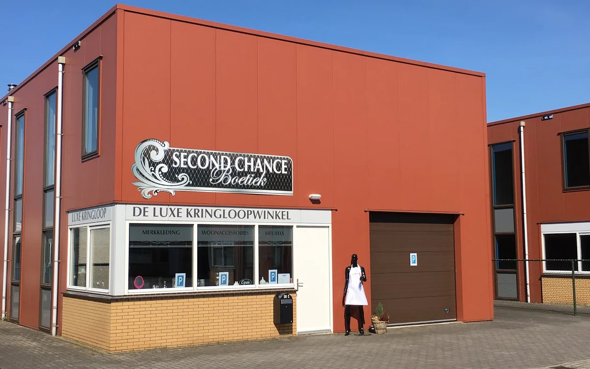 Rode bakstenen gevel van kringloopwinkel Second Chance Boetiek met witte winkelruit en bruine deur, persoon loopt langs