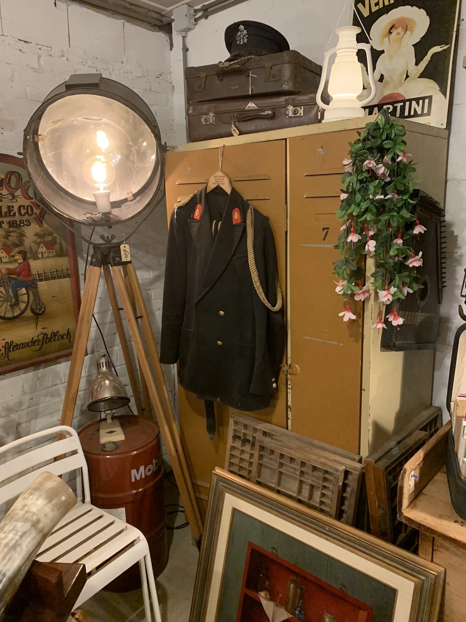 Interieur van kringloopwinkel met vintage lamp op statief, zwarte blazer op hanger, bloemenslinger en houten meubilair
