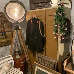 Interieur van kringloopwinkel met vintage lamp op statief, zwarte blazer op hanger, bloemenslinger en houten meubilair