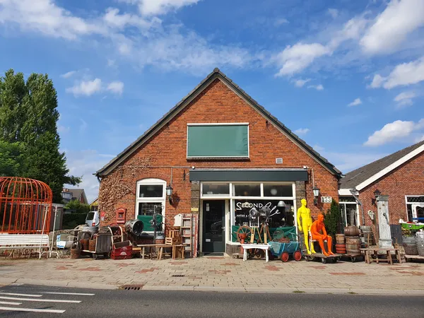 Voorgevel van kringloopwinkel Second Life Brocante, een rood bakstenen gebouw met groene raamluiken en uitgestalde spullen.