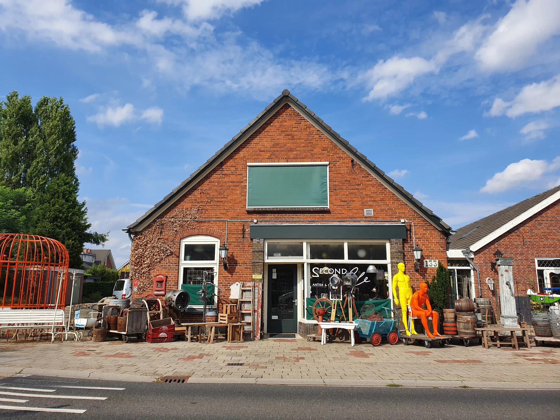 Voorgevel van kringloopwinkel Second Life Brocante, een rood bakstenen gebouw met groene raamluiken en uitgestalde spullen.