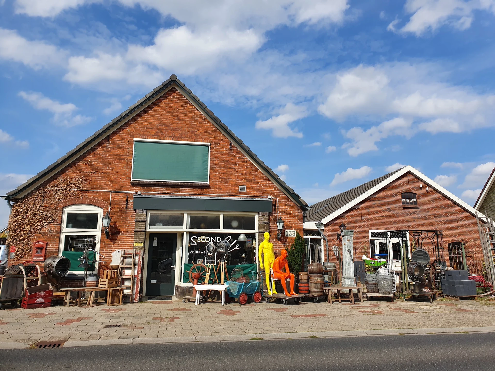 Rode bakstenen kringloopwinkel met groene ramen, uitgestalde spullen op het terras en bij de ingang