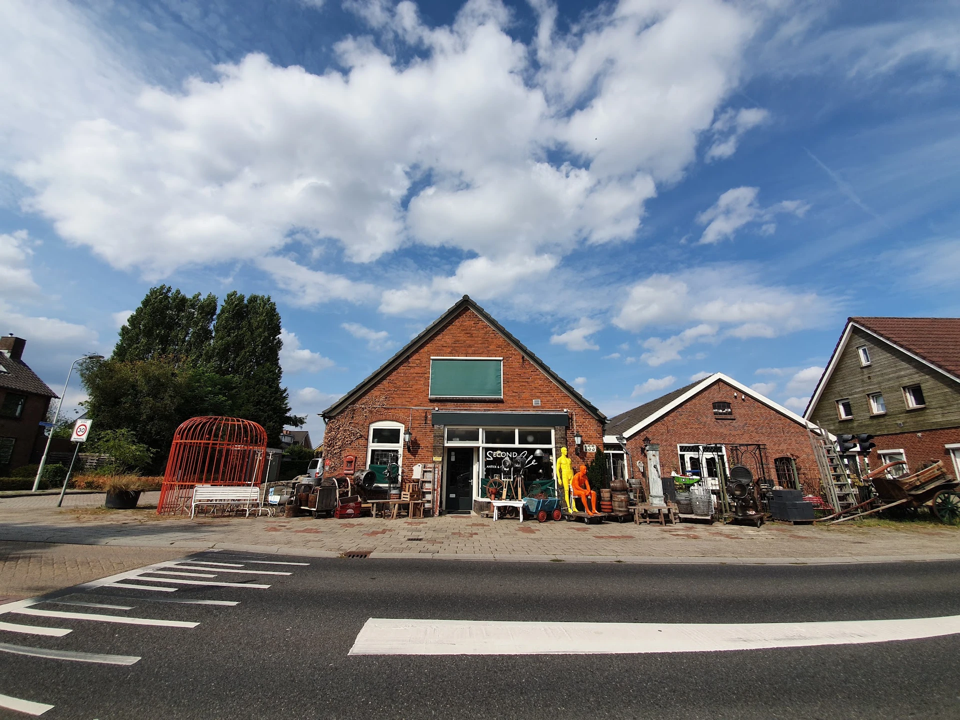 Voorgevel van bruine bakstenen kringloopwinkel met groene luiken, rode container links, grijze deur centraal, helder weer.