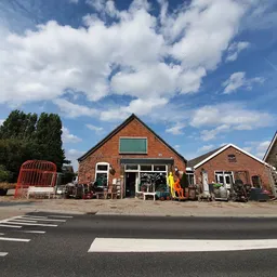 Voorgevel van bruine bakstenen kringloopwinkel met groene luiken, rode container links, grijze deur centraal, helder weer.