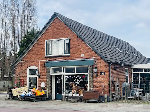 Bakstenen voorgevel van kringloopwinkel Second Life Brocante met groene winkelruit, uitgestalde spullen op straat en terras e