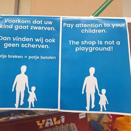 Twee blauwe borden met waarschuwingsteksten in Nederlands en Engels over toezicht op kinderen in de winkel