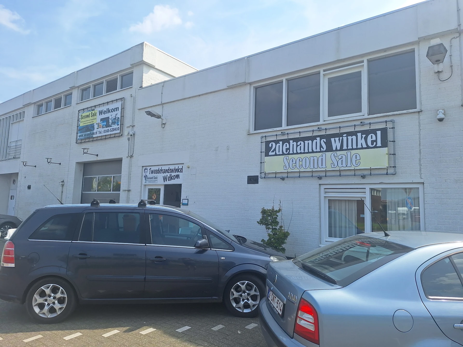 Witte gevel van kringloopwinkel Second Sale met winkelnaam op bord, parkeerplaats met twee auto's ervoor