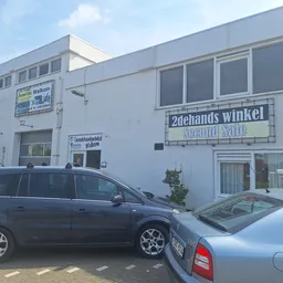 Witte gevel van kringloopwinkel Second Sale met winkelnaam op bord, parkeerplaats met twee auto's ervoor