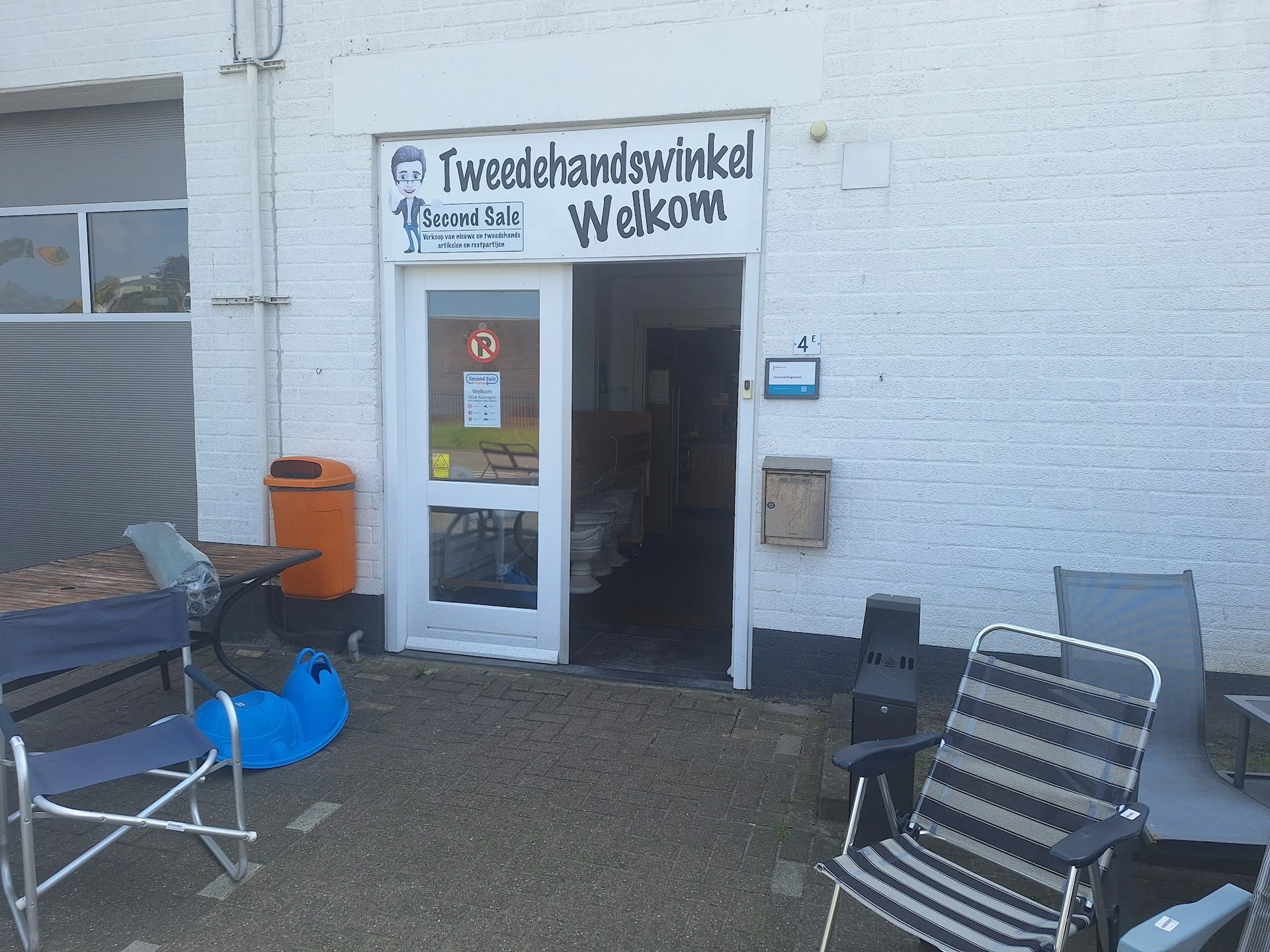 Witte gevel van Tweedehandswinkel Welkom met ingang, uithangbord en terras met stoelen
