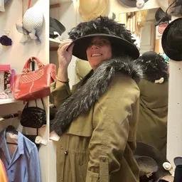 Vrouw in gele jas met zwarte bontje probeert zwarte hoed en roze handtas